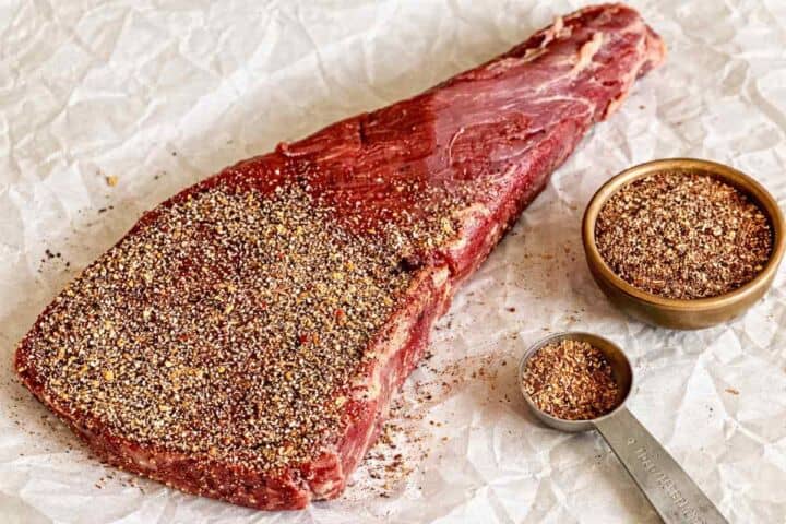 A raw steak covered in a coarse spice rub sits on parchment paper next to a metal tablespoon filled with seasoning and a small bowl of the same spice blend.