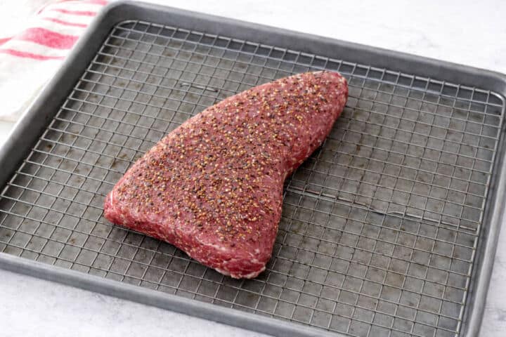A raw, seasoned tri-tip steak rests on a wire rack over a baking sheet. The surface of the meat is coated with cracked black pepper and spices. A red-striped towel is partially visible in the corner.