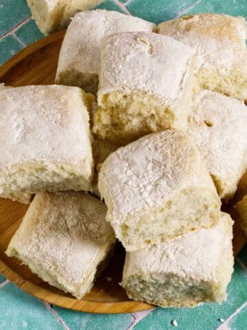 A wooden plate holds several fluffy, square bread rolls dusted with flour, set on a green tiled surface.