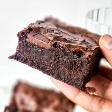 A hand holds a thick, fudgy chocolate brownie with a crinkly top and visible chocolate chunks, against a blurred background.