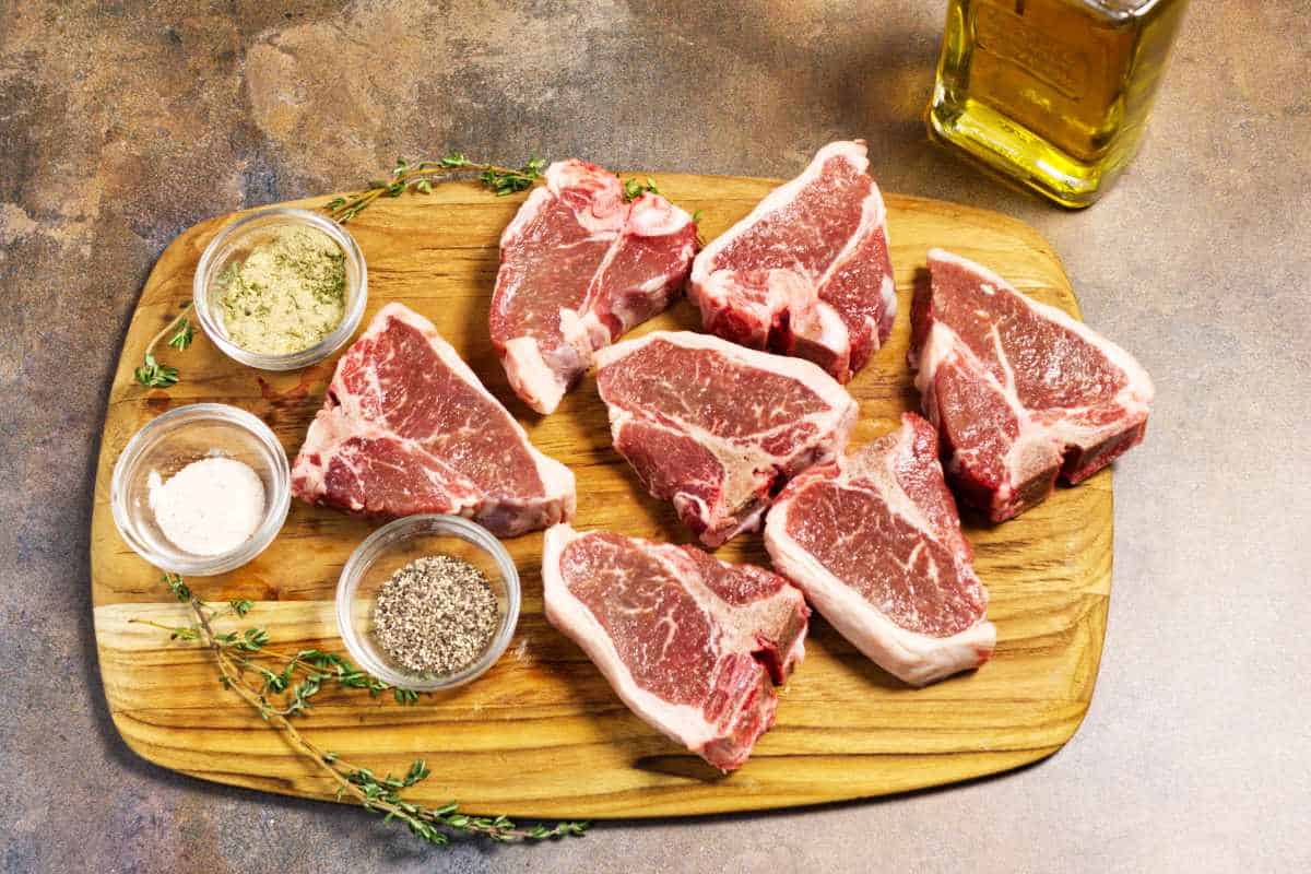 Raw lamb loin chops on a board with rosemary, garlic powder, salt, pepper, butter, and Italian dressing.