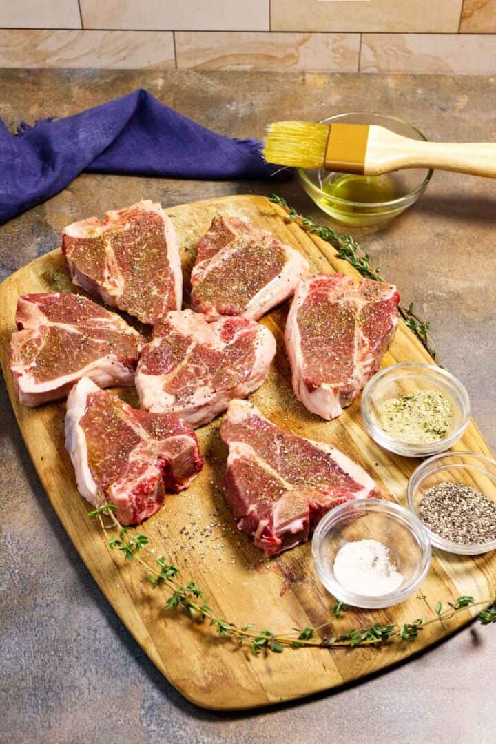 Raw lamb loin chops seasoned on a cutting board with salt, pepper, garlic powder, rosemary, and butter.