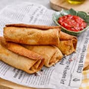 Four crispy, golden-brown chimichangas stacked on newspaper with a small bowl of red salsa for dipping beside them.