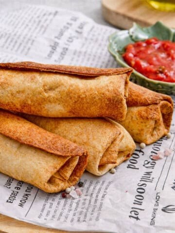 Four crispy, golden-brown chimichangas stacked on newspaper with a small bowl of red salsa for dipping beside them.