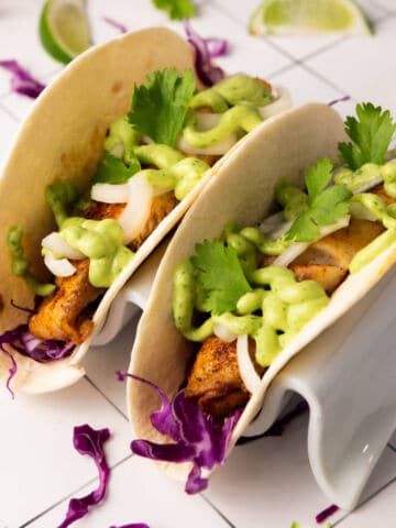 Two tacos filled with grilled fish, purple cabbage, sliced onions, cilantro, and creamy avocado crema, served in a white taco holder on a tiled surface with lime wedges and cilantro scattered around.
