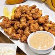A plate of crispy fried chicken bites with two lemon wedges, served alongside a bowl of creamy dipping sauce. The food is arranged on a white rectangular platter on a wooden surface.
