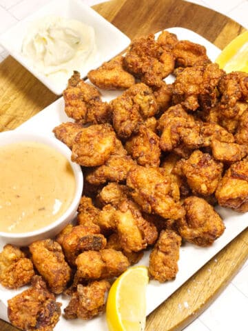 A white rectangular plate of golden-brown fried chicken bites, served with lemon wedges and two dipping sauces—one creamy white and one light orange—on a wooden board.