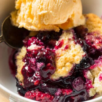A close-up of blackberry cobbler topped with a scoop of vanilla ice cream in a bowl. The cobbler has a golden crumbly crust with juicy, dark purple blackberry filling.