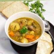 A bowl of Irish lamb stew with chunks of lamb, potatoes, and carrots sits on a black plate with buttered bread slices and fresh parsley on the side. The text “IRISH LAMB STEW” appears at the top.