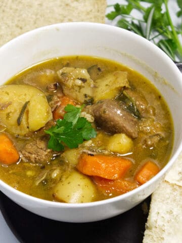 A bowl of hearty beef stew with chunks of beef, potatoes, carrots, and herbs, garnished with parsley. Slices of buttered bread and fresh parsley are on the side, with a spoon nearby.