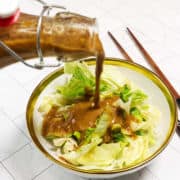 A glass bottle pours brown salad dressing over chopped iceberg lettuce in a white bowl with a gold rim. Chopsticks lie beside the bowl on a white, tiled surface.