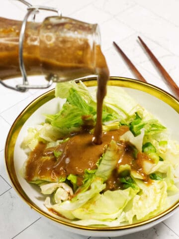 A glass bottle pours brown salad dressing over chopped iceberg lettuce in a white bowl with a gold rim. Chopsticks lie beside the bowl on a white, tiled surface.