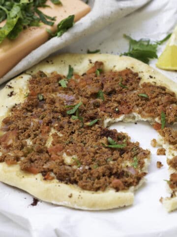 A round flatbread topped with crumbled spiced mixture, diced tomatoes, and herbs sits on white paper. A slice is missing. Nearby are a lemon wedge, fresh herbs, and a peach-colored block on a light cloth.