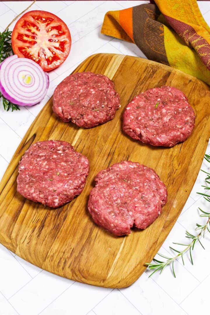 Four raw lamb burger patties on a cutting board with tomato and red onion nearby.