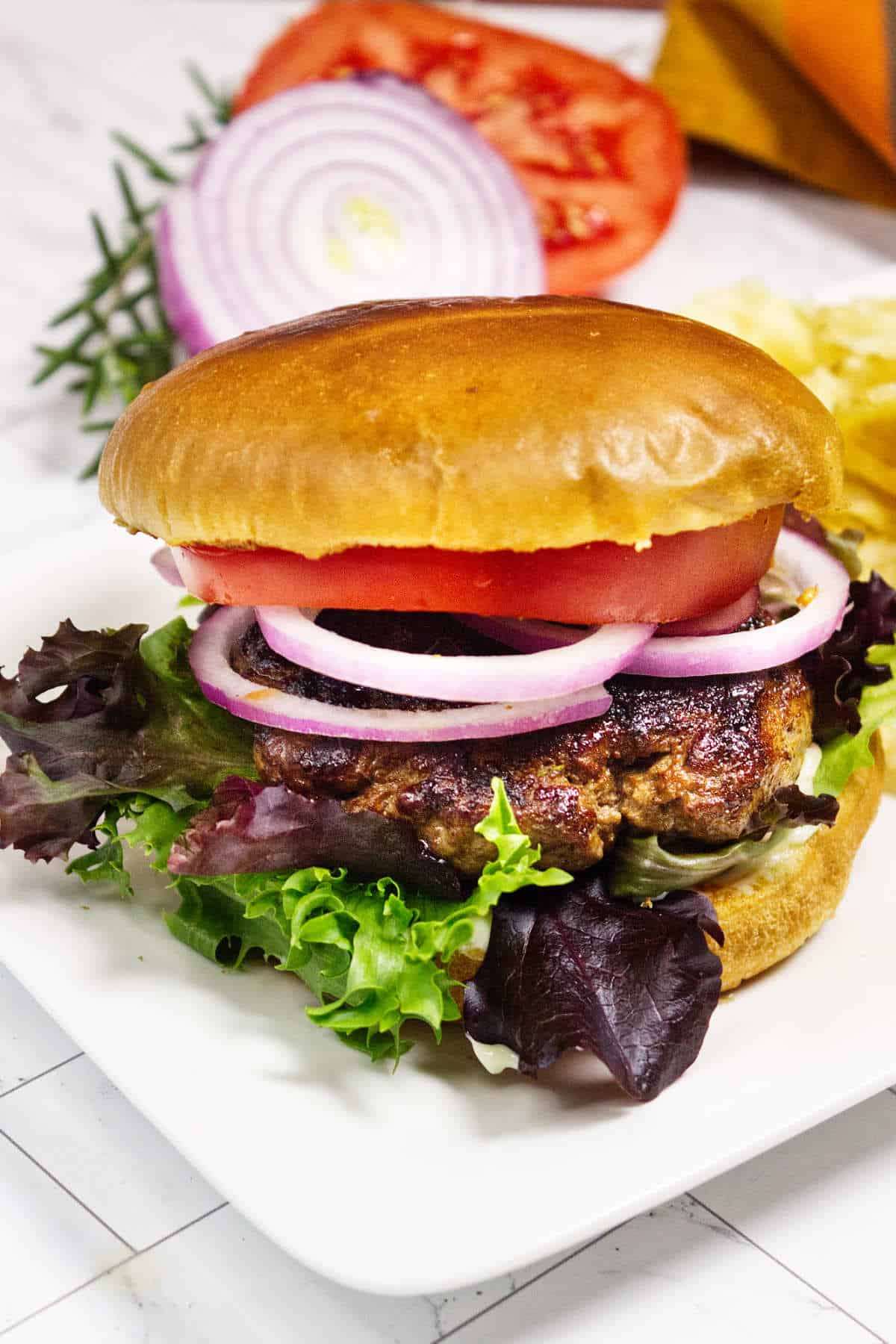 Lamb burger on a white plate with lettuce, tomato, red onion, and potato chips.