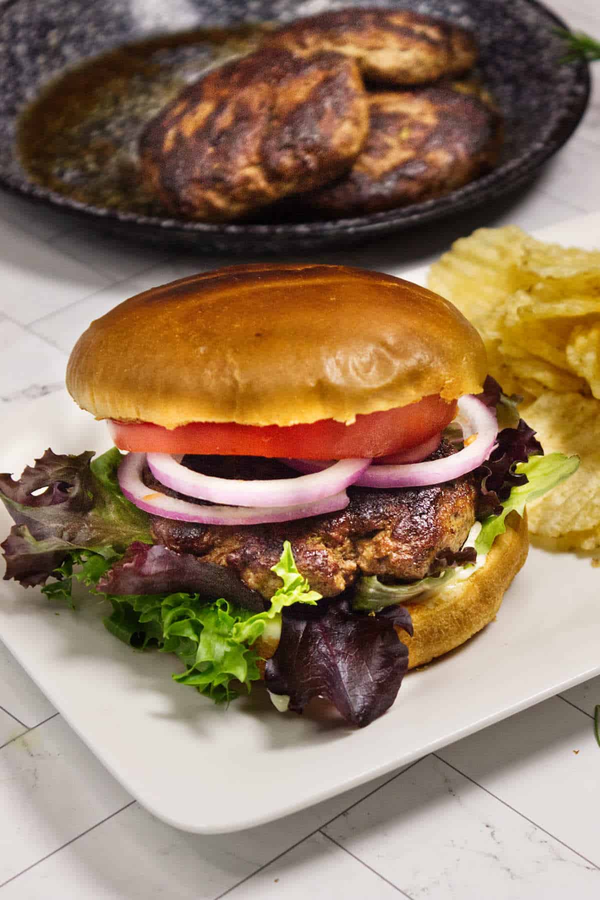 Lamb burger with lettuce, tomato, red onion, and potato chips on a white plate.
