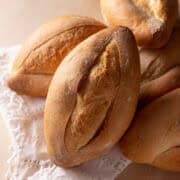 A close-up of several oval-shaped, crusty bolillo rolls placed on a white, lace-trimmed cloth, showcasing their golden-brown, crisp crusts and rustic appearance.
