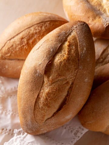 A close-up of several oval-shaped, crusty bolillo rolls placed on a white, lace-trimmed cloth, showcasing their golden-brown, crisp crusts and rustic appearance.