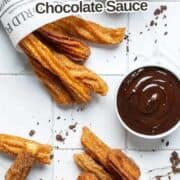 Two paper cones filled with Mexican churros are on a white tiled surface, next to a bowl of chocolate sauce. Some churros and chocolate crumbs are scattered around. Text reads: “Mexican Churros with Chocolate Sauce.”.