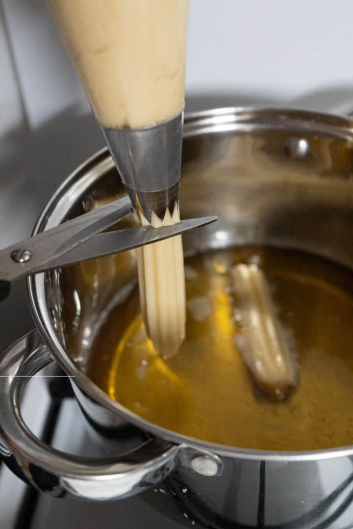 Piping churro dough into hot oil using a star tip and cutting strips.