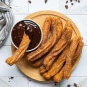 A wooden plate with several cinnamon-sugar churros next to a small bowl of chocolate sauce; one churro is dipped in the sauce. A striped cloth and chocolate bits are on the white tiled surface.