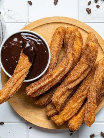 A wooden plate with several cinnamon-sugar churros next to a small bowl of chocolate sauce; one churro is dipped in the sauce. A striped cloth and chocolate bits are on the white tiled surface.