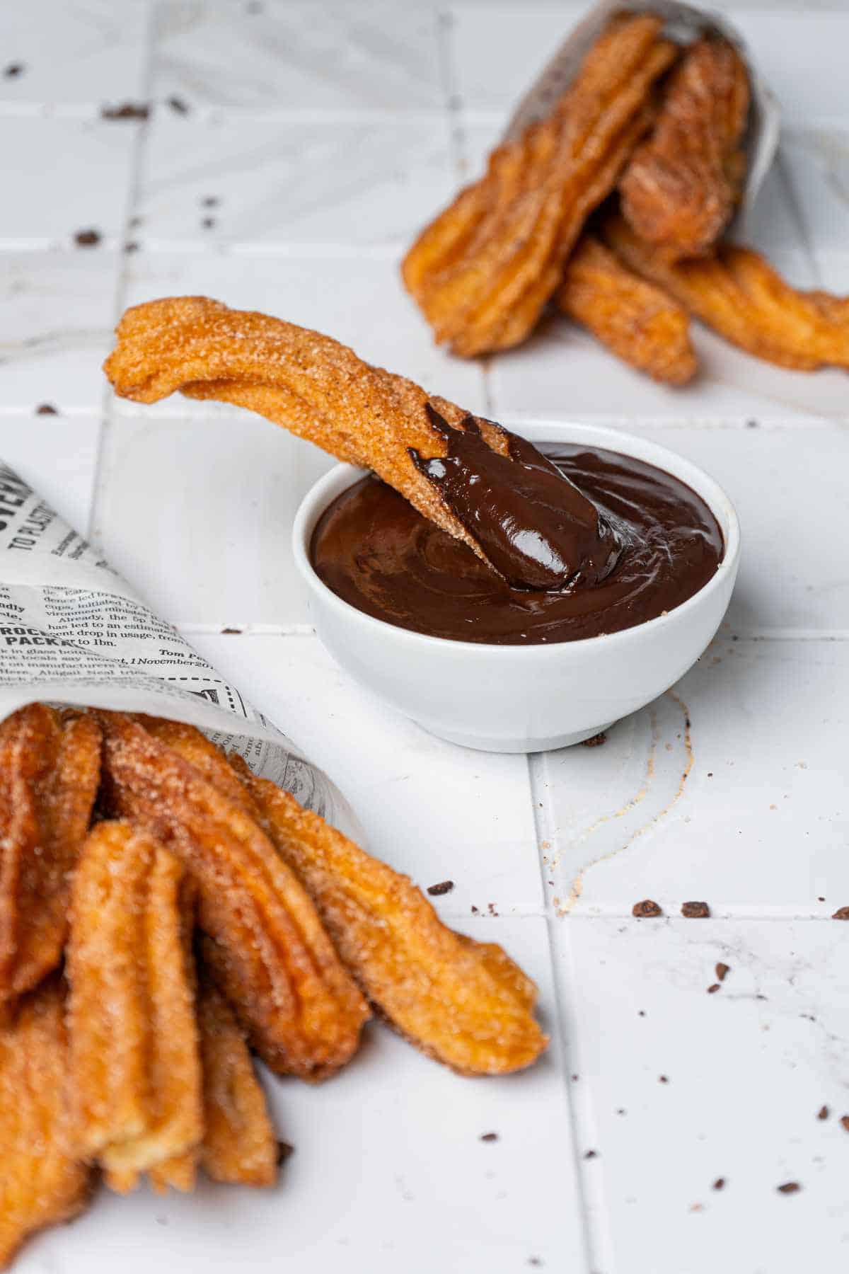 A bowl of chocolate sauce with a churro dipped in it is on a white tiled surface. More churros, covered in sugar, are scattered around and wrapped in paper nearby.