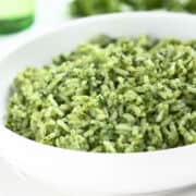A close-up of a white bowl filled with green rice, likely prepared with herbs or spinach, giving the rice a vibrant green color. The background is softly blurred.