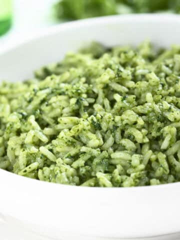 A close-up of a white bowl filled with green rice, likely prepared with herbs or spinach, giving the rice a vibrant green color. The background is softly blurred.