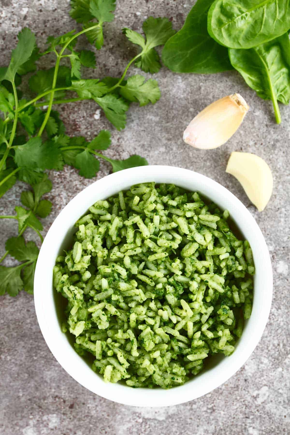 Arroz verde in a white bowl with cilantro, garlic, and greens nearby.