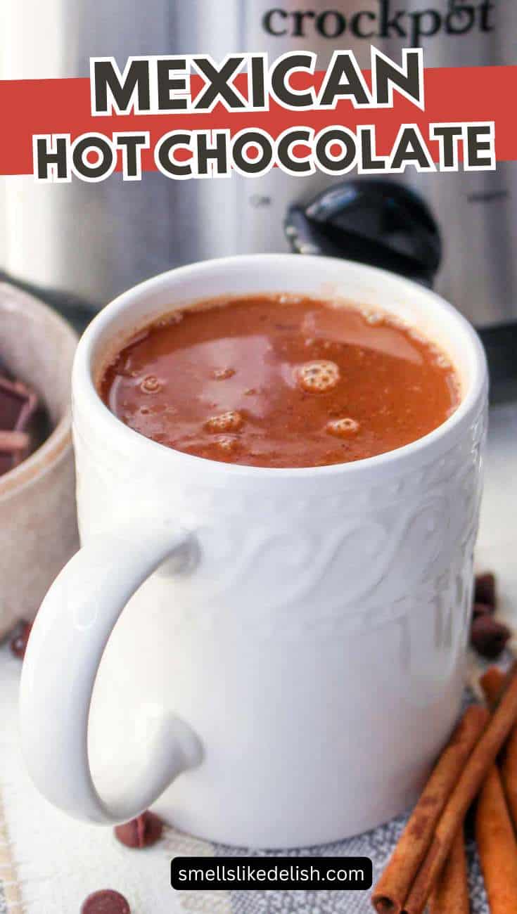 Mexican hot chocolate in a white mug with a slow cooker, cinnamon sticks, and chocolate pieces nearby.