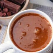 A white mug filled with creamy Mexican hot chocolate sits on a table. Beside it are chocolate bars, chocolate chips, and cinnamon sticks. The words "Mexican Hot Chocolate" appear at the top.