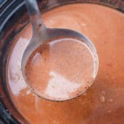A metal ladle filled with smooth, creamy Mexican Hot Chocolate held above a pot containing more of the same liquid.