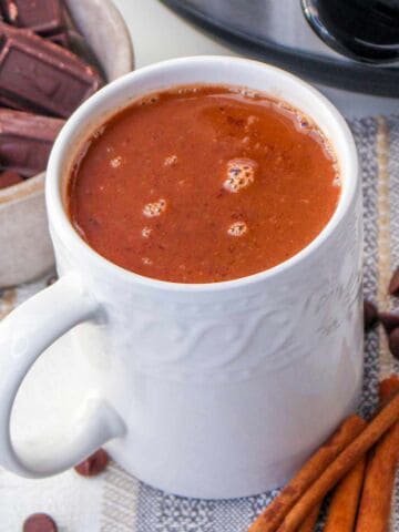 A white mug filled with hot chocolate sits on a striped cloth, surrounded by cinnamon sticks, chocolate bars, chocolate chips, and part of a slow cooker.