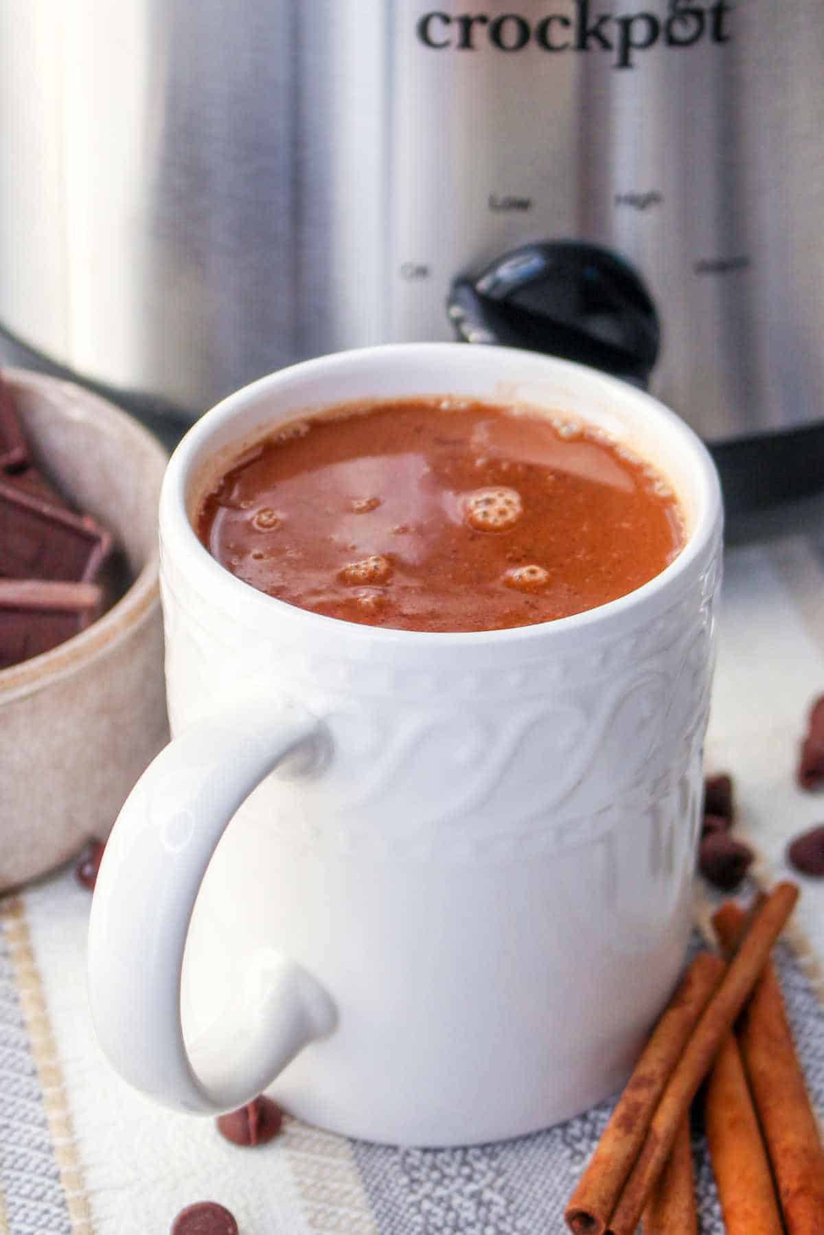 White mug filled with Mexican hot chocolate in front of a Crockpot with cinnamon sticks and chocolate pieces nearby.
