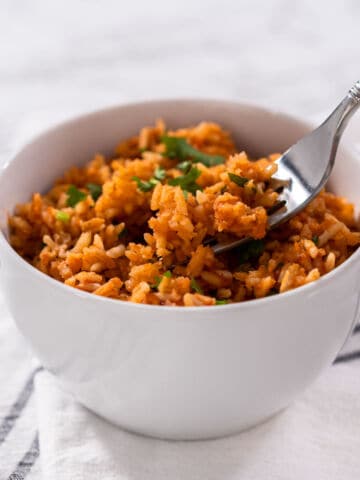 A white bowl filled with Mexican rice garnished with chopped herbs, with a fork taking a portion from the bowl. The bowl sits on a white cloth with black stripes.