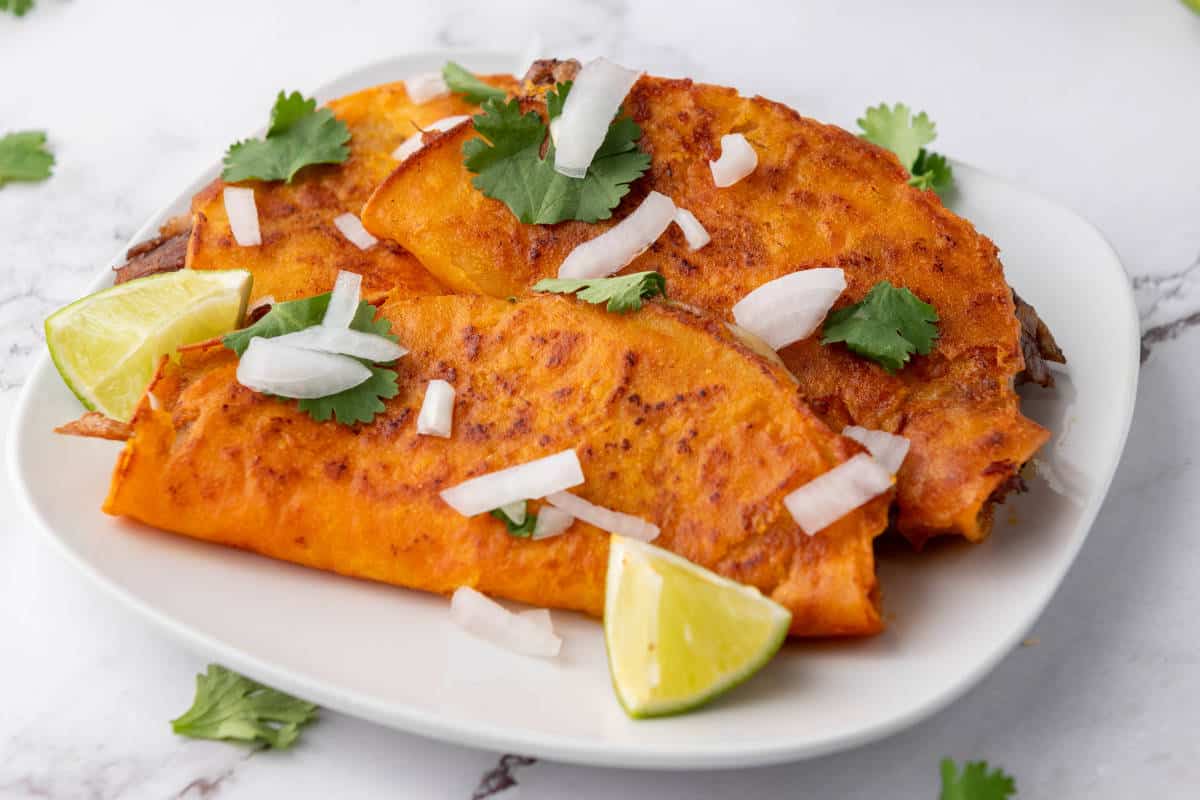 Quesabirria tacos garnished with chopped onions, cilantro, and lime wedges on a white plate.