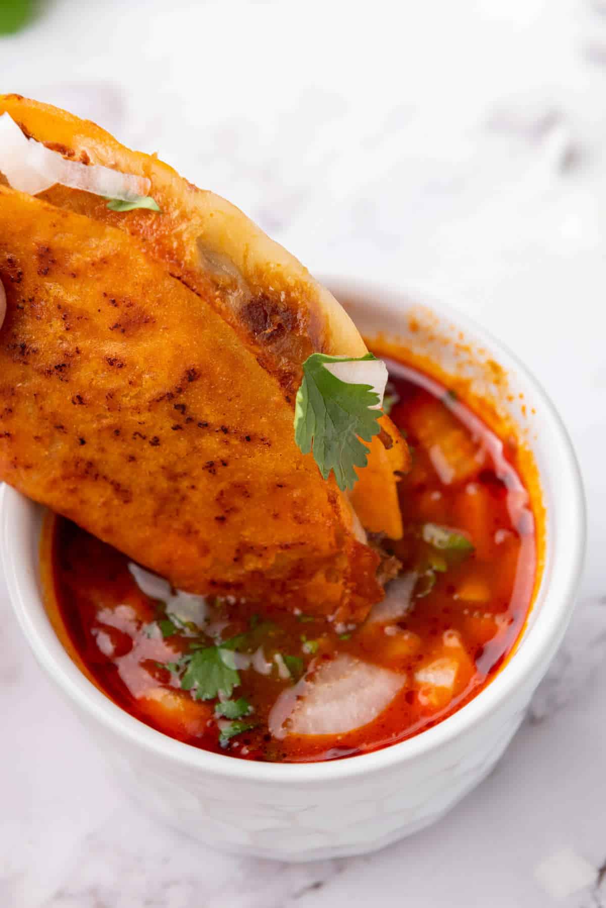 A crispy quesabirria taco dipped into a cup of red consome with chopped onions and cilantro.