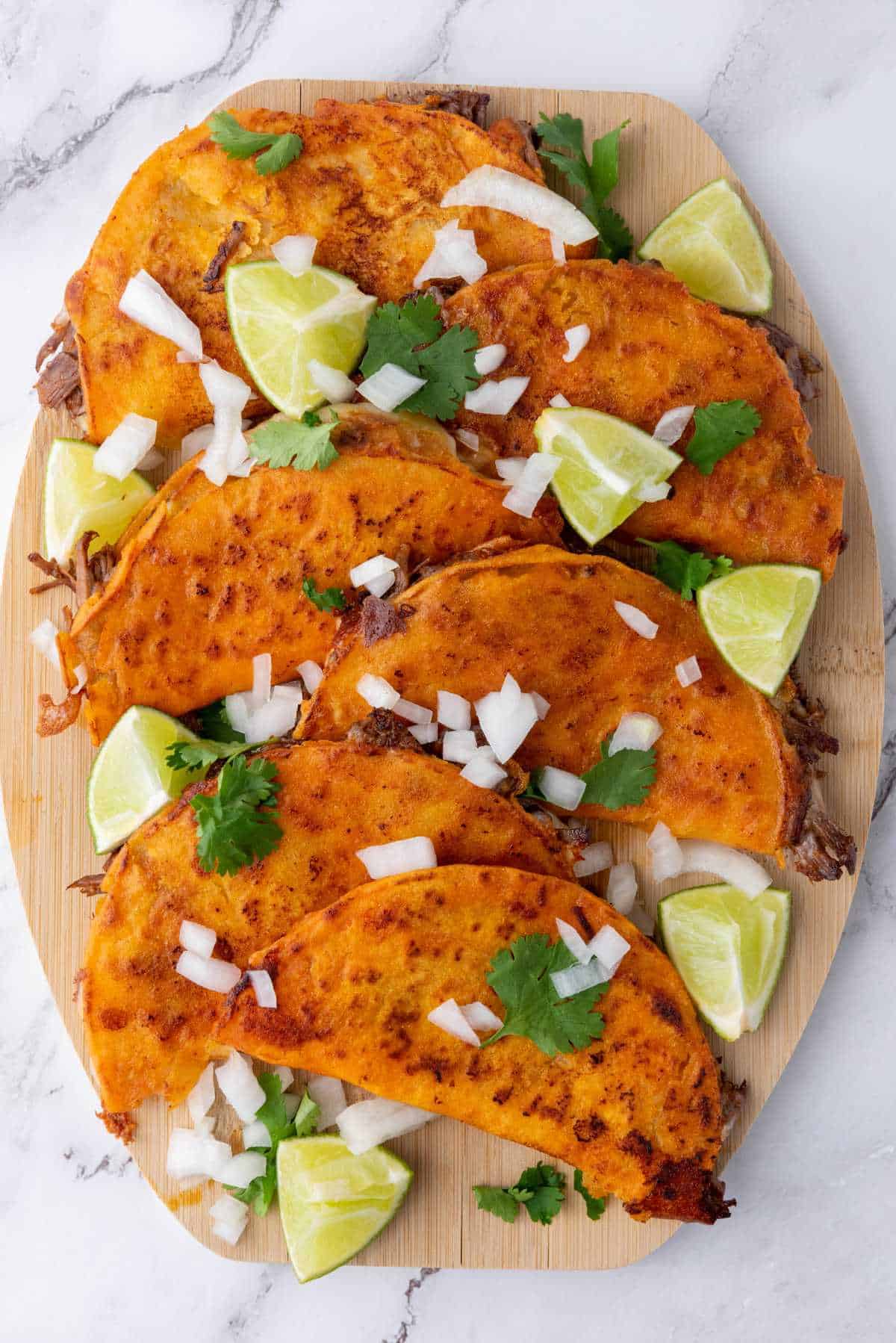 A board of crispy quesabirria tacos topped with onions and cilantro served with lime wedges.