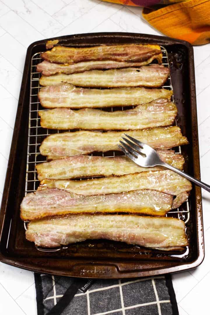 Partially cooked bacon on a rack over a sheet pan with a fork used to flip the strips.