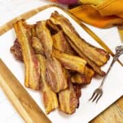 A white plate piled with crispy, cooked bacon strips sits on a wooden board. A decorative silver fork rests on the plate, and a yellow and red cloth is visible in the background.