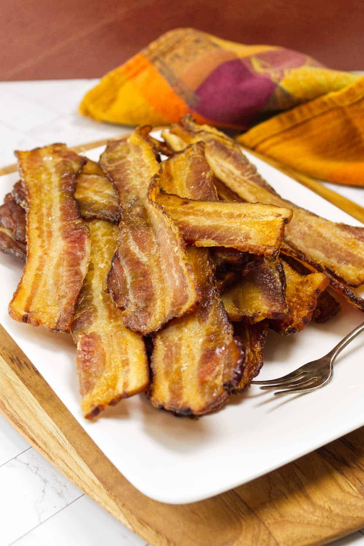 Crispy oven baked bacon strips on a white serving plate with a fork and cloth napkin.