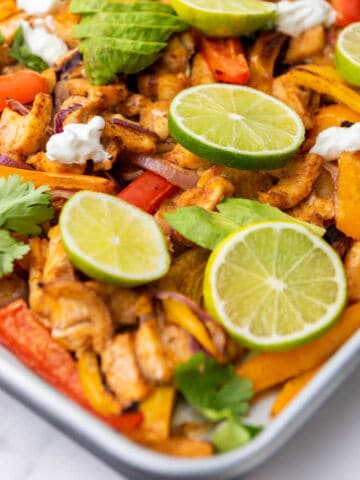 Colorful sheet pan chicken fajitas with sliced red and yellow bell peppers, red onion, cooked chicken strips, fresh cilantro, lime slices, avocado, and dollops of sour cream on a white tray.