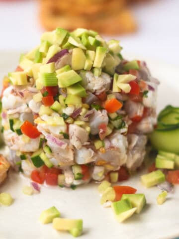 A close-up of a seafood ceviche salad with diced avocado, tomato, red onion, and cucumber, garnished with cucumber ribbons and served on a white plate.