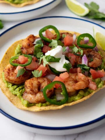 A crispy tortilla topped with guacamole, grilled shrimp, sliced jalapeños, diced tomatoes, red onions, cilantro, and a dollop of sour cream, with a lime wedge on the side.