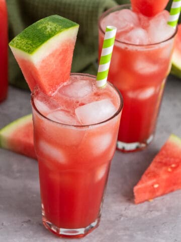 Two tall glasses of watermelon juice with ice cubes, each garnished with a watermelon wedge and a green-striped straw. Slices of watermelon are placed on a light gray surface nearby.