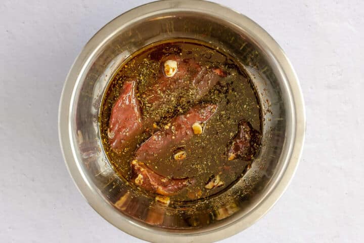 Steak marinating in a stainless steel bowl.