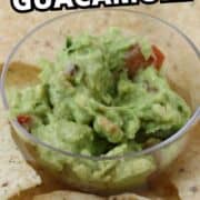 A clear bowl of guacamole with tomato pieces sits on a plate surrounded by tortilla chips.