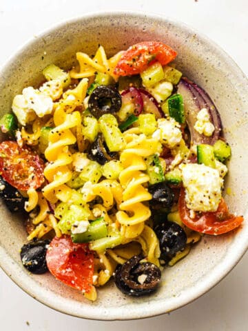 Bowl of spiral pasta salad with cherry tomatoes, black olives, cucumber, red onion, feta cheese, and light dressing.