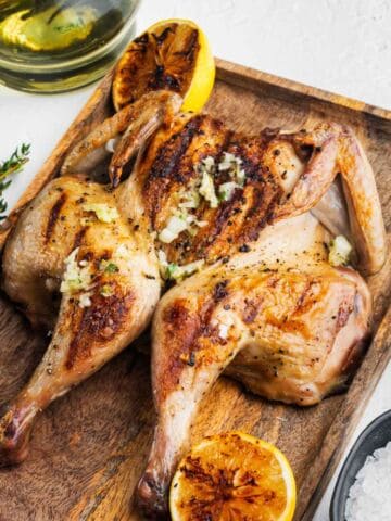 Roasted spatchcock chicken with garlic, herbs, and charred lemon on a tray, surrounded by salt, peppercorns, herbs, and wine.